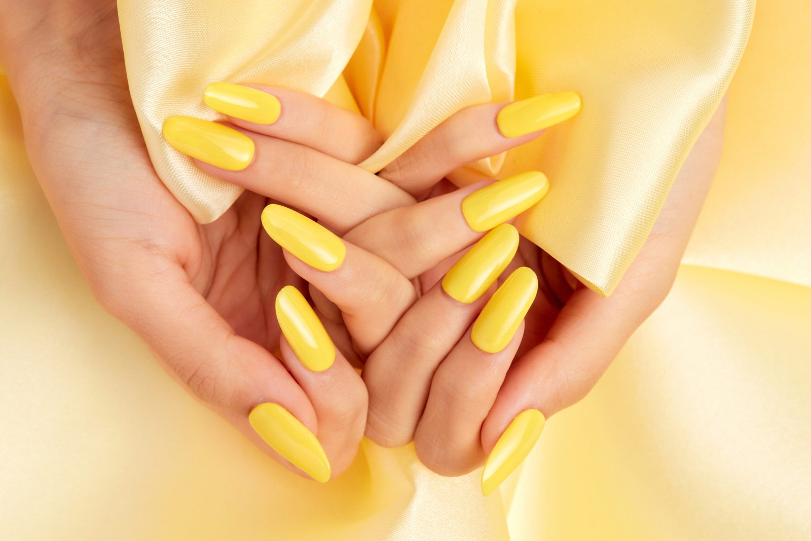 A closeup shot of a female's hands with yellow nail polish on a yellow silk fabric